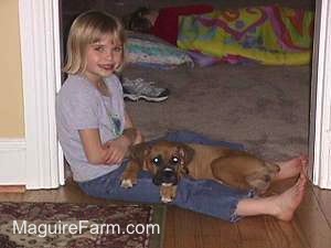 A Blonde Girl is sitting in a doorway and A Tan Boxer Puppy is laying in her lap