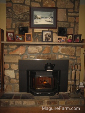 There is a black tea pot on top of the newly inserted wood burning stove. There is a lit fire. The fireplace mantel has family pictures on it. There is a paiting of a farm house in the snow with a blue border above the mantel.