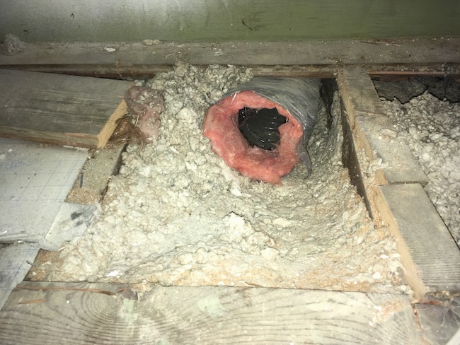 A large black flex duct with pink fiberglass exposed poking out from the gray blown-in insulation where the floor boards were removed in an attic.