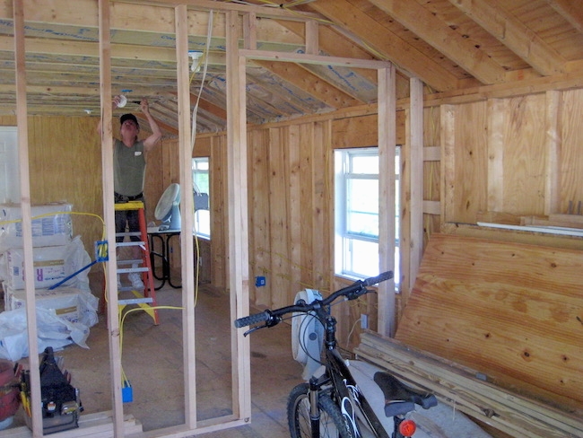 A studded room with a bike parked and in the back room is a man on a step ladder putting up light fixtures and piles of fiberglass batts in white plastic on the floor.