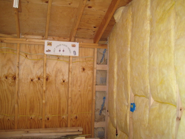 A studded room with yellow fiberglass lined on one side and the empty studs on the other. There is a paper sign hanging on the wall that says 'get your food here'