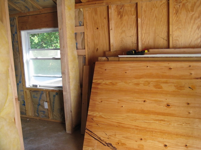 A studded wall with plywood leaning against the wall. There is an open window in the next room with fiberglass lining the wall and ceiling around the window.