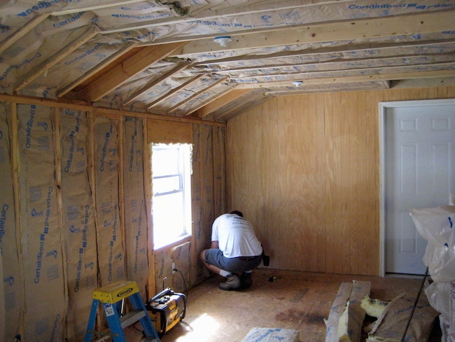 A man in a room bending down working on a wall that is lined with fiberglass batts