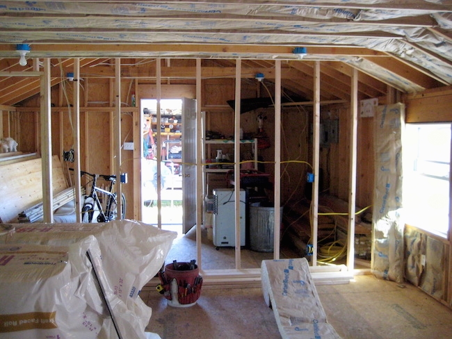 A room with the studs showing and fiberglass batts lining the walls and rolls of the spun glass on the floor awaiting instulation. There is a kids bicycle in the back of the room.