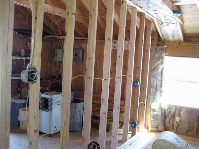 An unfinished room with the studs showing and fiberglass batts lining the walls and ceiling. There is a dehumidifier in the back room.
