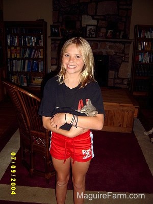 A girl standing inside a living room holding a little gray kitten with a smile on her face. She is wearing red shorts and a black shirt and there are book shelves behind her.