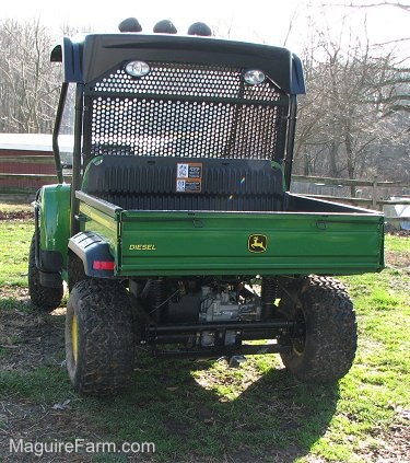 The back of a green and yellow John Deere HPX 4x4 Gator parked in the grass with a split rail fence and a chicken coop in front of it.