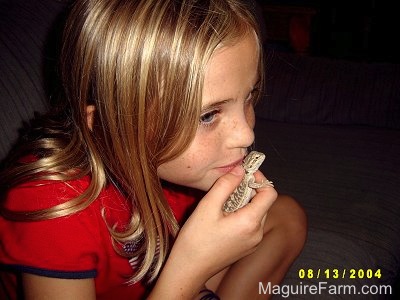 Close Up - A blonde haired girl with a red shirt on is kissing a young bearded dragon