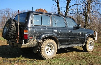 The back of a jacked up black 1996 Toyota Land Cruiser with mud on the large tires and a built in jack next to the spare tire.
