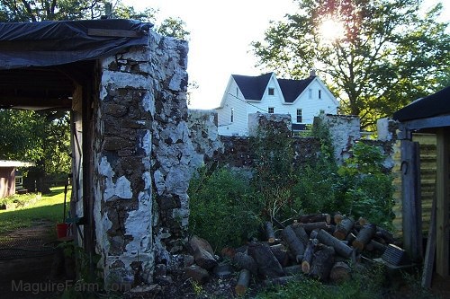 The stone walls of an old barn with a white farm house in the background