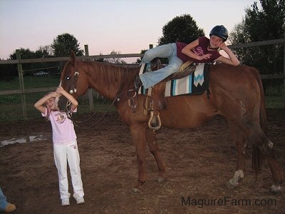 A girl in a burgundy shirt is laying across the top of the back of a brown horse and a second girl in a pink shirt is hugging the horses head.