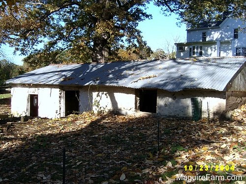 An old stone spring house showing an open doorway and two open windows. Just up the hill from it is an old white farm house.