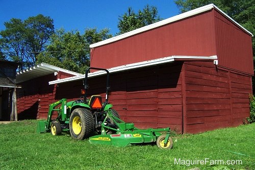 A John Deere 5065E Compact Tractor and Front End Loader with MX-7 cutting deck is parked behind a classic red barn
