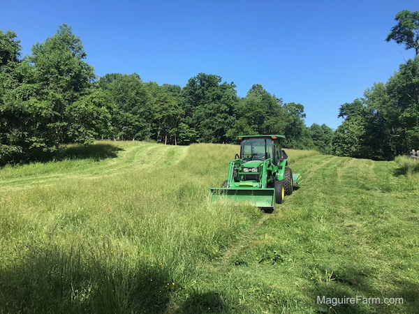 John Deere 5065E Compact Tractor and Front End Loader with a cutting deck cutting a feild with a line of trees behind it